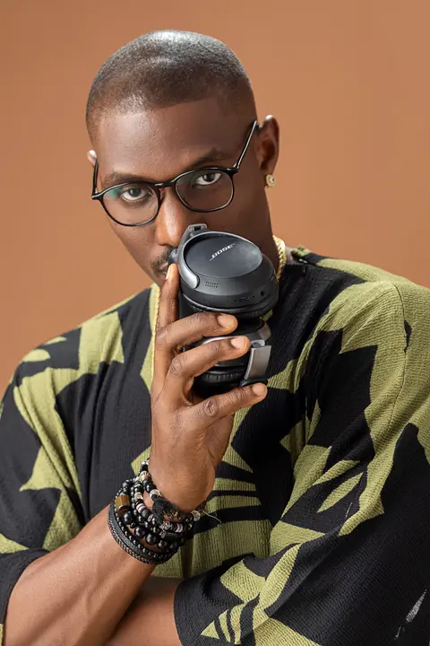 A man in glasses and a green and black print shirt sips from a black cup against tan.
