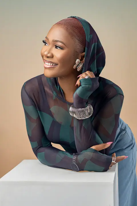 A smiling woman in a patterned headwrap leans on a white plinth.