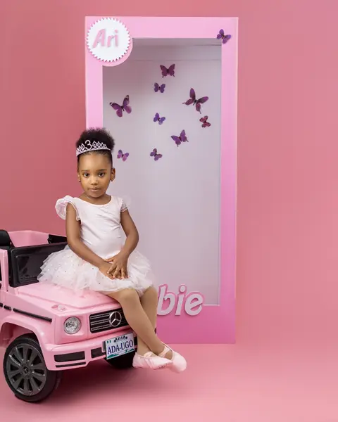 A little girl in a tiara sits in a pink toy car beside a doll-box set.