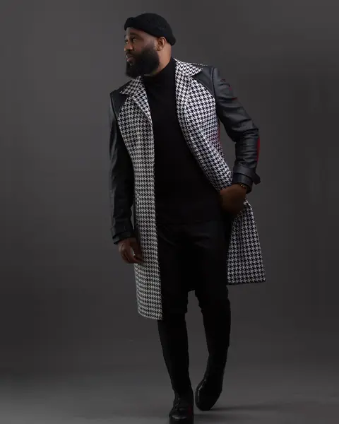 A bearded man in a houndstooth and leather coat walks against a grey backdrop.