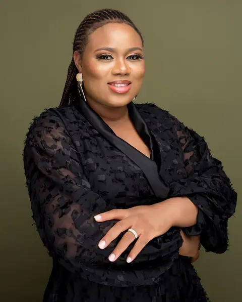 A woman with braided hair in black lace, arms folded, against olive.