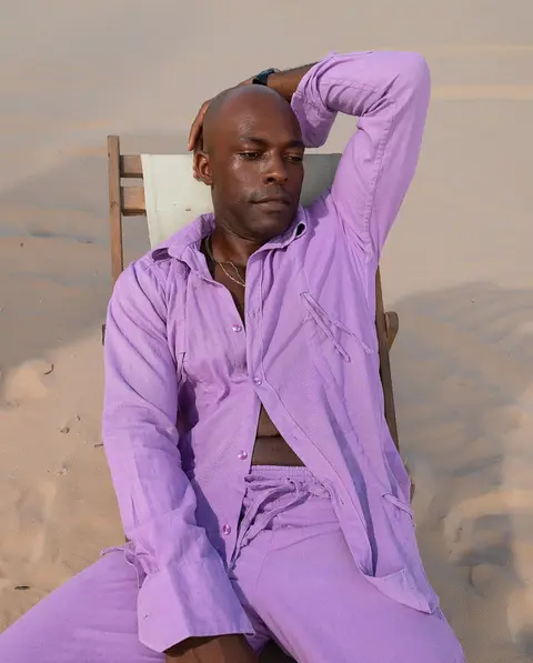 A man in a lilac shirt and trousers stretches back in a chair on the sand.