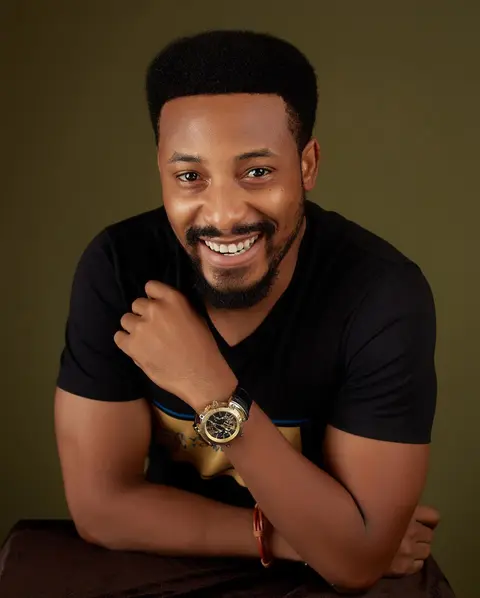A smiling man in a black tee and gold watch leans on his arm against olive.