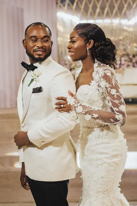 A groom in a white tuxedo and a bride in a lace mermaid gown pose close in the reception hall.