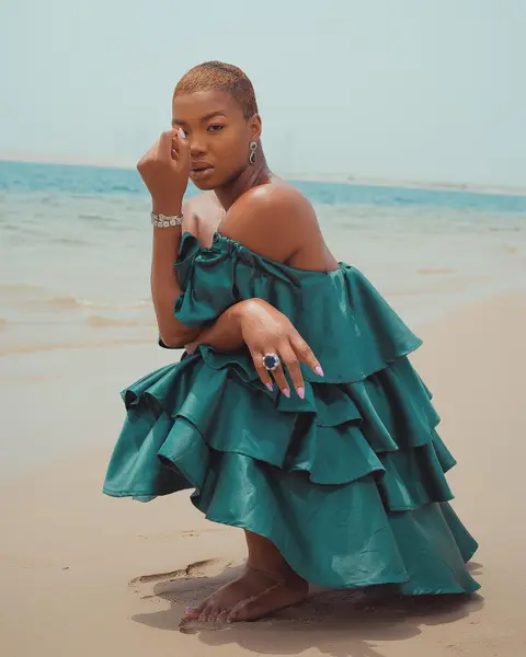 A woman in a tiered teal ruffle dress crouches on the sand by the sea.