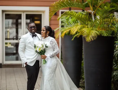The bride and groom smile together outside, she holding white calla lilies, he in a white dinner jacket.