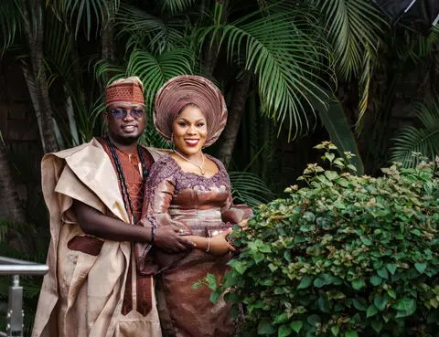 The couple in coordinated aso-oke and coral beads at their traditional ceremony, framed by palms.