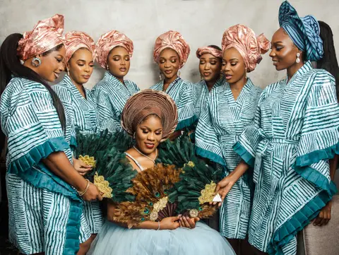 The bride in a peach gele surrounded by her bridesmaids in teal and white aso-oke holding green feather fans.