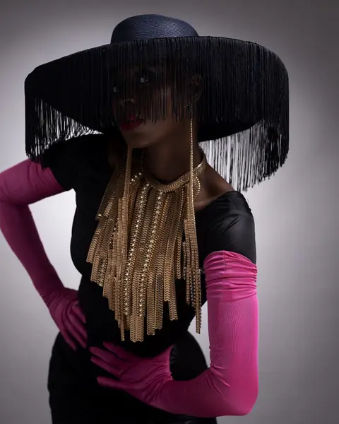 A model in a wide black fringed hat, gold fringe necklace and magenta opera gloves against grey.