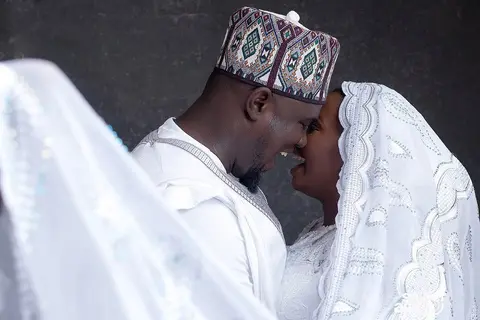 A couple in white, the groom in an embroidered cap, foreheads touching under a lace veil.